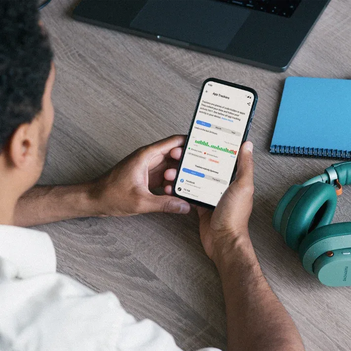 Person holding a Fairphone 5 showing an app trackers screen, on a wooden desk next to green headphones and a notebook